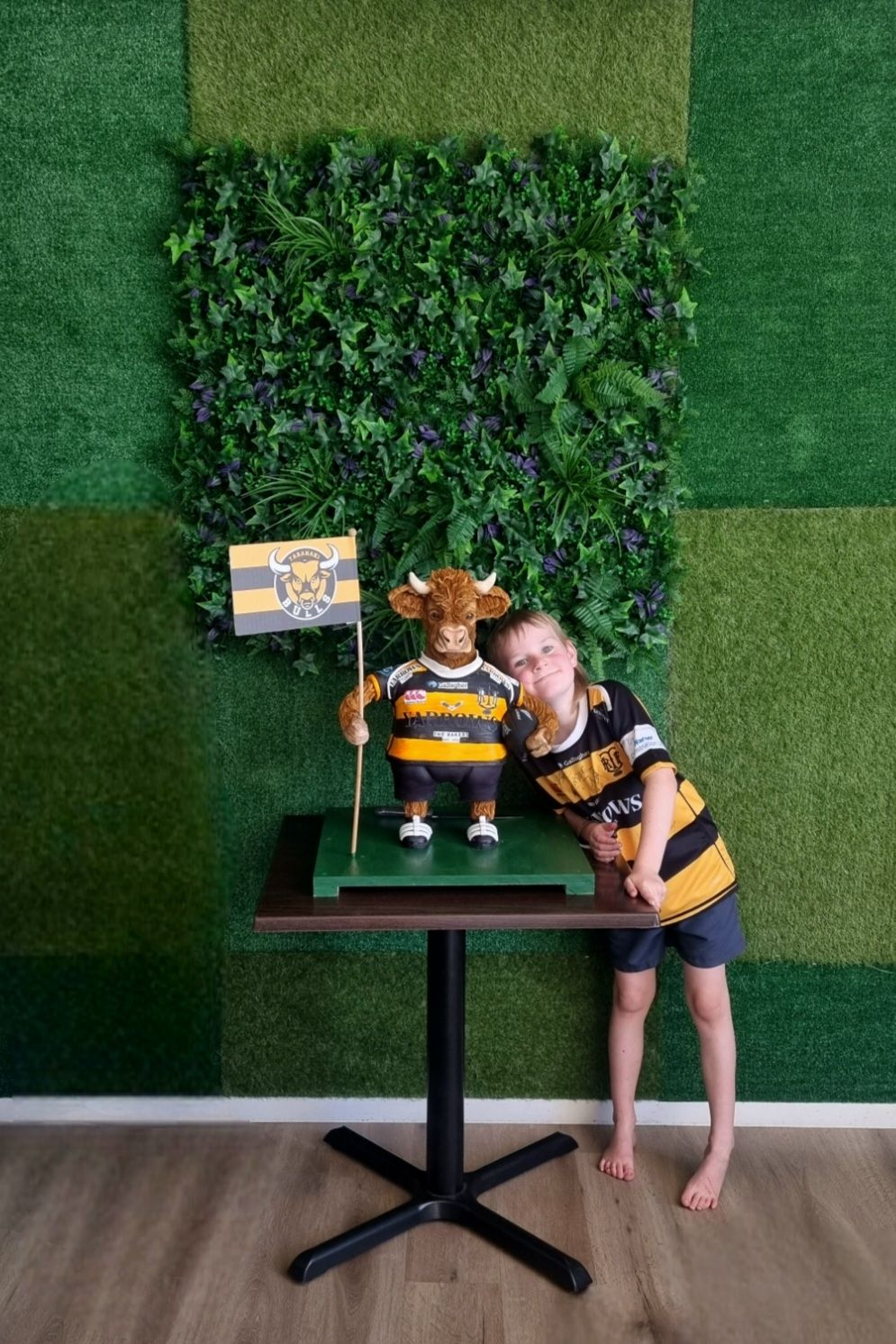 "Taranaki Bulls-themed sculpted bull cake displayed on a table alongside a smiling child wearing a matching rugby jersey, with a lush green foliage wall background, perfect for a sports-themed celebration."