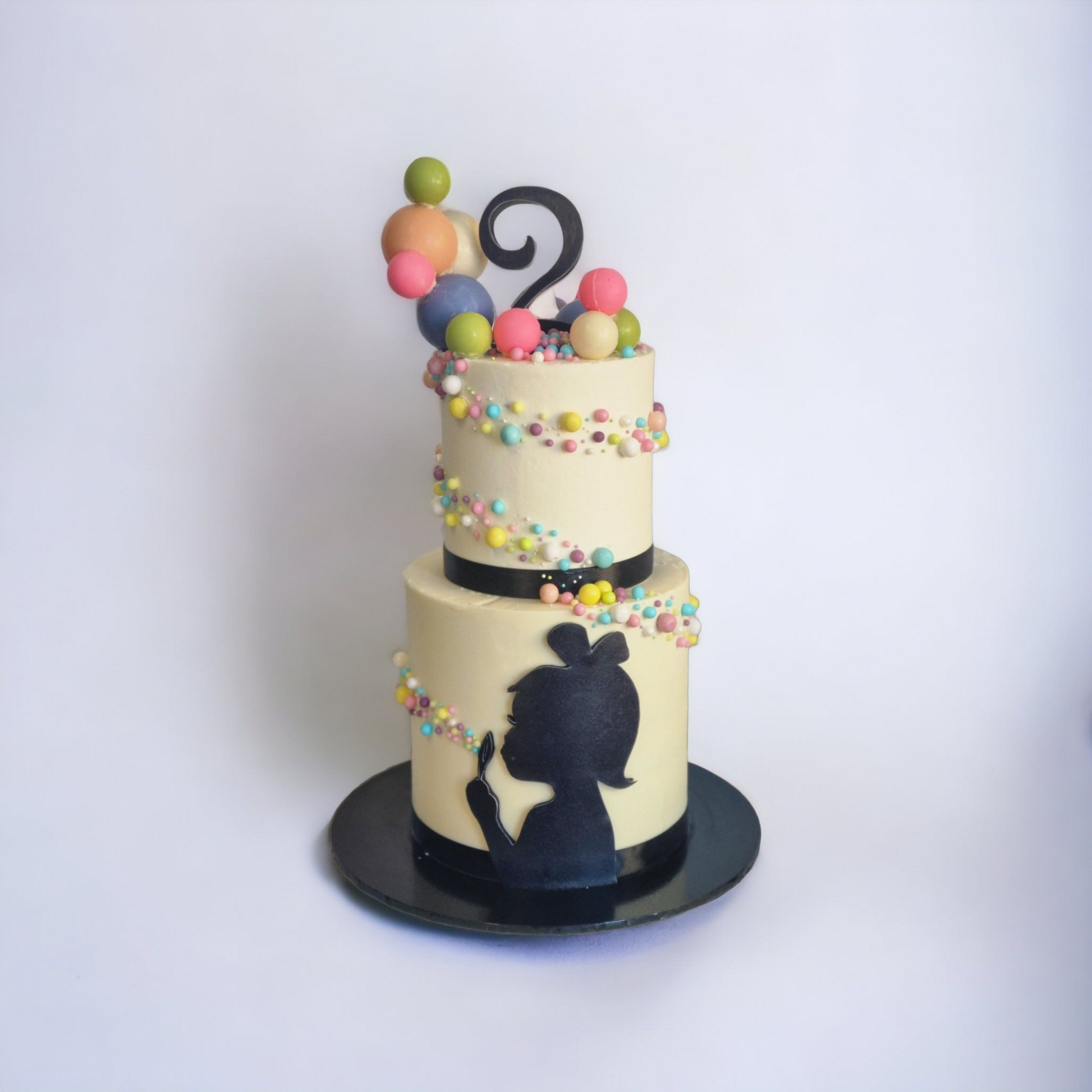 A two-tiered cake with a bubble-blowing theme, decorated with pastel-colored edible pearls and a silhouette of a child blowing bubbles on the bottom tier. A black ribbon wraps around the base of each tier, and a black number '2' topper sits on top, surrounded by colorful spherical decorations. The cake is placed on a glossy black cake board against a clean, white background.